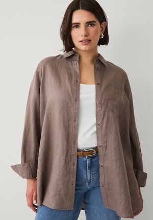 Vrouw met een open losse bruine overhemd met knopen over een wit shirt, gecombineerd met een blauwe spijkerbroek en een beige riem, staand tegen een effen achtergrond.