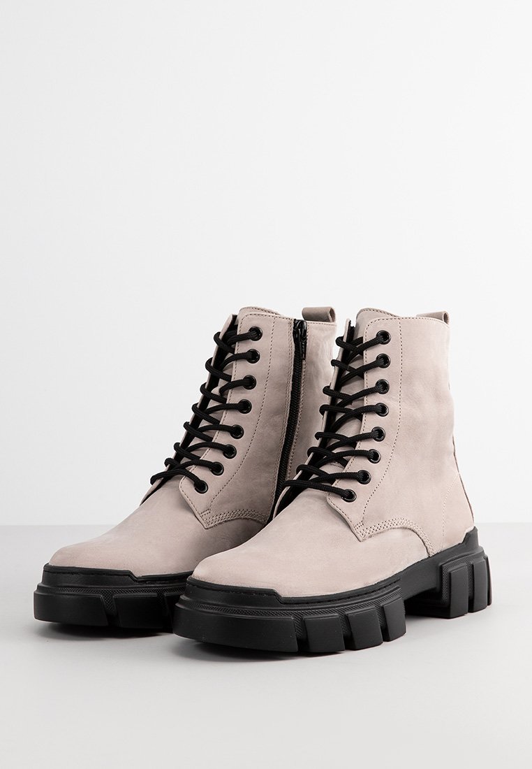 Botas de tobillo de ante beige con cordones negros, cremallera lateral y suelas negras gruesas. Presentan una textura suave y un diseño sencillo.