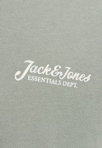 Tejido verde texturizado con el logo en blanco de "Jack & Jones" y el texto "ESSENTIALS DEPT." exhibido de manera prominente.