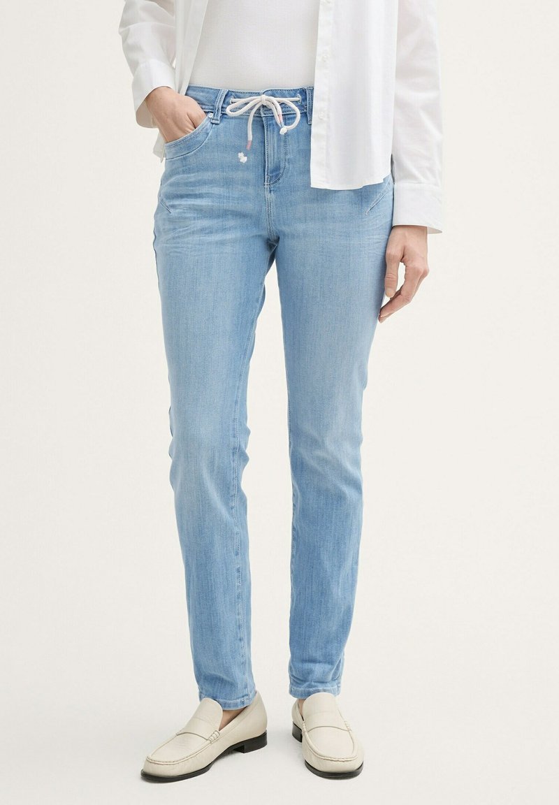 Světle modré džíny z denimu s páskem na stahování, slim fit a předními kapsami. V kombinaci s bílou košilí na knoflíky a béžovými mokasíny.