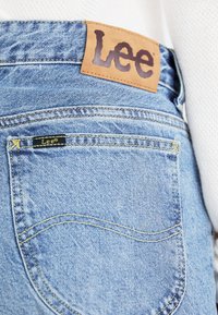 Blue denim jeans featuring a leather waistband label with "Lee" logo, yellow stitching, and back pocket detailing. Smooth texture.