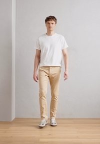 Hvit bomulls T-skjorte, beige slim-fit bukser og grå joggesko, stående på et tregulv mot en lys grå vegg.