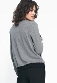 Kobieta z kręconymi ciemnymi włosami, ubrana w dopasowaną szaro-czarną marynarkę w wzór chevron oraz czarne spodnie, spoglądająca przez ramię.