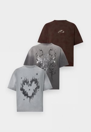En mand og en kvinde står side om side iført matchende brune grafiske t-shirts; kvinden i patchwork denim nederdel, manden i beige bukser, begge i hvide sneakers.