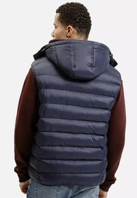 Mężczyzna w granatowej, pikowanej kamizelce z kapturem nosi bordową koszulę z długim rękawem, stoi tyłem na tle białym.