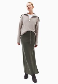 Beige zip-up sweatshirt, olivengrøn lang nederdel med lynlås foran, parret med mørke ankelstøvler. Glat stoftekstur og tætsiddende design.