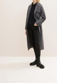 Cappotto grigio lungo su una blusa nera, abbinato a jeans neri slim-fit e stivali neri robusti. Tessuto liscio con una silhouette sartoriale.