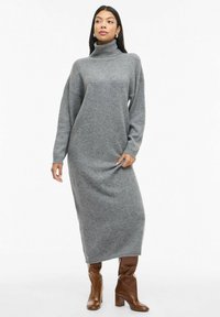 Abito in maglia grigio a collo alto con spalle scese, maniche lunghe e lunghezza alla caviglia, abbinato a stivali con tacco marroni. Texture liscia.