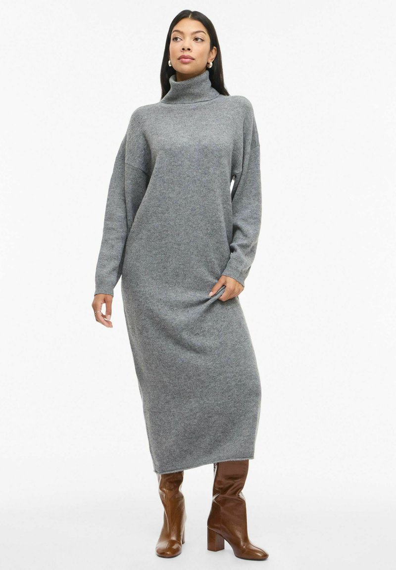 Abito in maglia grigio a collo alto con spalle scese, maniche lunghe e lunghezza alla caviglia, abbinato a stivali con tacco marroni. Texture liscia.