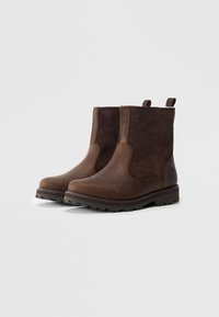 Botas de tobillo de cuero marrón con un acabado suave, paneles laterales elásticos y suela de goma texturizada. Cuenta con lengüetas para facilitar el uso.
