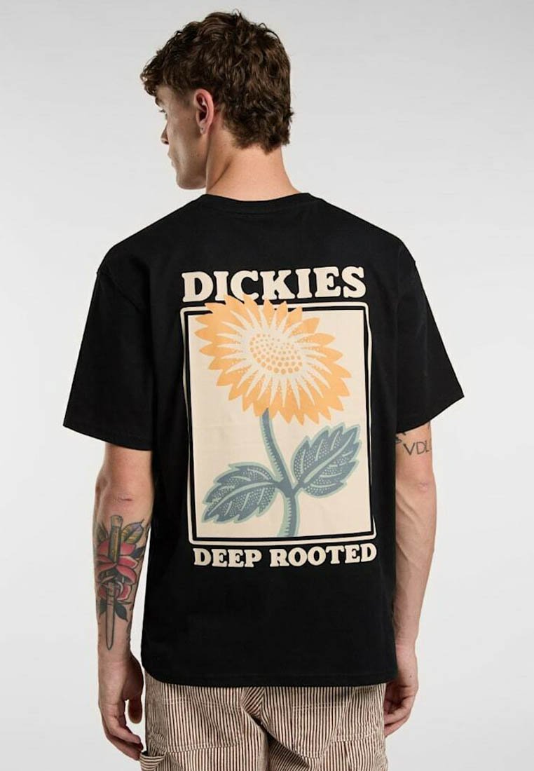 Ung mand med kort krøllet hår iført en sort T-shirt med en stor solsikkegrafik og teksten "DICKIES DEEP ROOTED" på bagsiden.