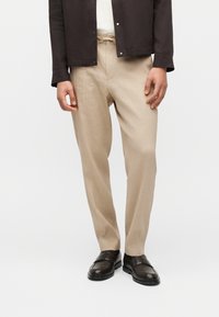 Άνδρας που φοράει μπεζ παντελόνι με κορδόνι στη μέση, λευκό πουκάμισο, σκούρο καφέ σακάκι και μαύρα δερμάτινα μοκασίνια, στέκεται μπροστά σε ένα απλό φόντο.