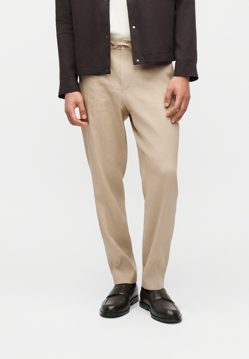 Άνδρας που φοράει μπεζ παντελόνι με κορδόνι στη μέση, λευκό πουκάμισο, σκούρο καφέ σακάκι και μαύρα δερμάτινα μοκασίνια, στέκεται μπροστά σε ένα απλό φόντο.