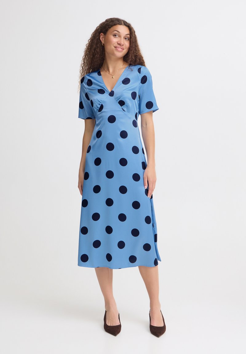 Femme aux cheveux bouclés portant une robe midi bleue à pois noirs et des escarpins noirs pointus, debout devant un fond clair uni.