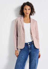 Blazer rose clair avec un design ajusté, fermeture à un bouton et revers crantés, associé à un haut blanc et un jean bleu.