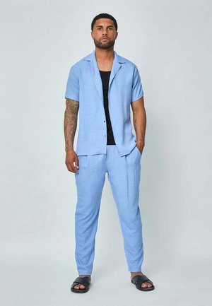 Chemise à manches courtes bleu clair à boutons, associée à un pantalon léger assorti, avec une texture subtile. Portée avec des sandales noires à semelle plate.