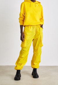 Personne portant un sweat à capuche jaune vif et un pantalon cargo assorti avec des grosses bottes noires, debout devant un fond blanc.