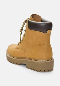 Beige Wildleder-Ankle-Boot mit einem braunen Lederpatch am Schaft, goldenen Ösen und einer dicken, robusten Sohle für besseren Halt.