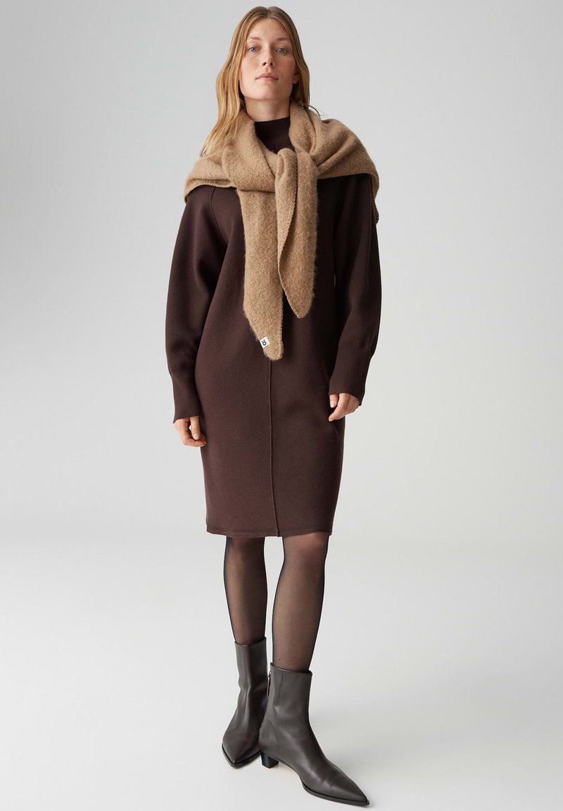 Braunes Strickkleid mit langen Ärmeln, geripptem Halsausschnitt und Schlitz am Rücken. Hellbrauner, weicher Schal, der über die Schultern drapiert ist. Schwarze Stiefeletten.