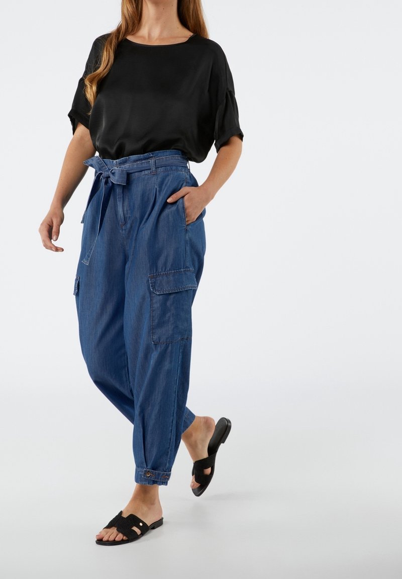 Blusa de satén negra con mangas cortas, combinada con pantalones cargo de mezclilla sueltos que incluyen cinturón, bolsillos laterales y puños enrollados. Sandalias planas negras.