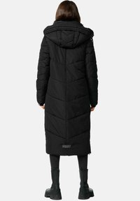 Sort lang pufferjakke med hætte, med chevron-syning, et tekstureret stof og et logo nederst. Båret over mørke bukser og støvler.