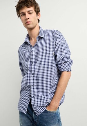 Junger Mann mit zerzausten Haaren, der ein blau-weiß kariertes Hemd und blaue Jeans trägt, steht mit einer Hand in der Tasche, neutraler Hintergrund.