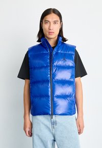 Gilet rembourré bleu avec col montant, motif matelassé horizontal, zip sur le devant et accent logo. Porté par-dessus une chemise noire avec un jean en denim clair.
