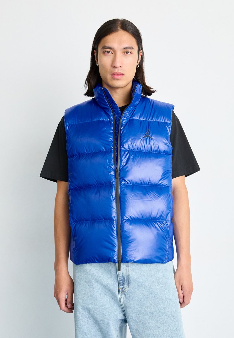 Gilet rembourré bleu avec col montant, motif matelassé horizontal, zip sur le devant et accent logo. Porté par-dessus une chemise noire avec un jean en denim clair.