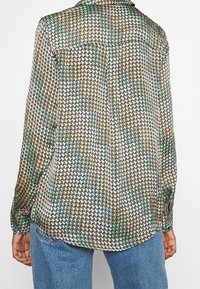 Long-sleeve blouse made of lightweight fabric featuring a houndstooth pattern in green, black, and yellow, with a curved hem and buttoned cuffs.