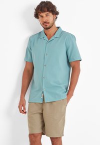 Light blue short-sleeve shirt with a textured pattern, featuring a open collar, front buttons, and a tag. Paired with beige shorts.