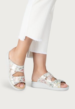 Feet wearing white floral open-toe slip-on sandals paired with white cropped pants with frayed hems.