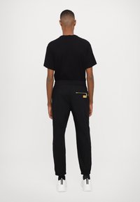 Black short-sleeve t-shirt and black joggers with elastic waistband. Joggers feature a zippered pocket with gold hardware. White sneakers.