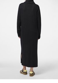 Lungo vestito nero a maglia con collo alto, spacco laterale e maniche lunghe. Il tessuto appare morbido e texture. Indossato con sneakers marroni e gialle.
