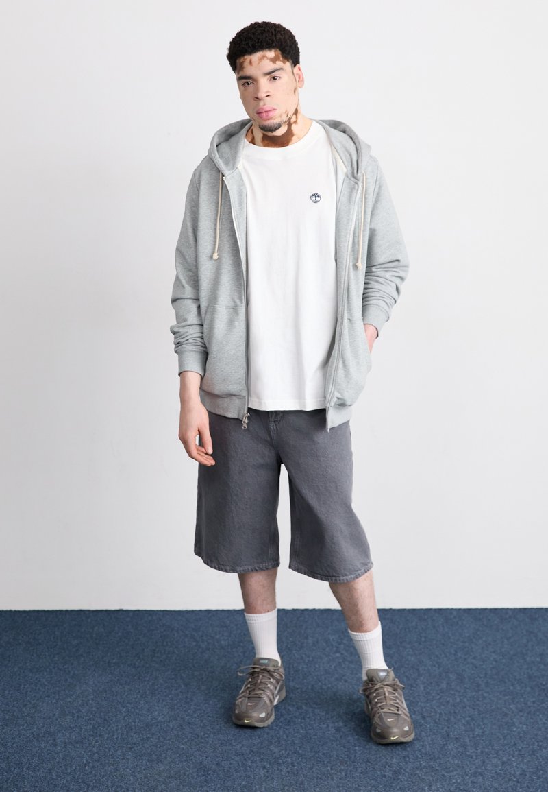 Sudadera gris con cremallera, camiseta blanca, pantalones cortos grises, zapatillas grises y negras, con calcetines blancos. Tejido suave, corte relajado, diseño casual.