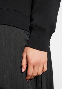 Schwarzer Pullover mit gerippten Bündchen und glatter Textur, kombiniert mit einem schwarzen Rock mit dezentem Glanz; die Hand ruht auf dem Kleidungsstück.