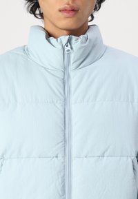 Light blue puffer jacket with a high collar, front zipper, quilted texture, and visible stitching detail.