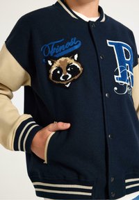 Marinelblauwe varsityjack met beige mouwen, gestreepte manchetten, geborduurde wasbeer en tekst; voorzien van drukknoopsluiting en zijzakken.