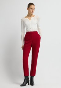 Weißes Langarmshirt mit vertikalen Streifen und Knöpfen, kombiniert mit hoch taillierten roten Hosen und schwarzen Stiefeletten.