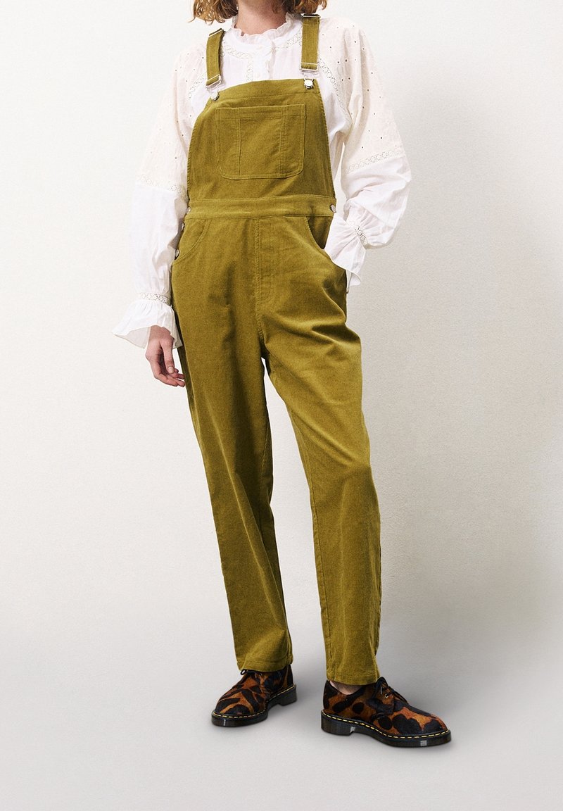 Olivengrønne kordsfløjlsbukser med justerbare stropper og forlomme, kombineret med en hvid bluse med flæseærmer og mønstrede sko.