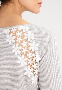 Top a rayas grises y blancas con un acento de encaje floral en la parte posterior, con un escote redondo y un corte ajustado.