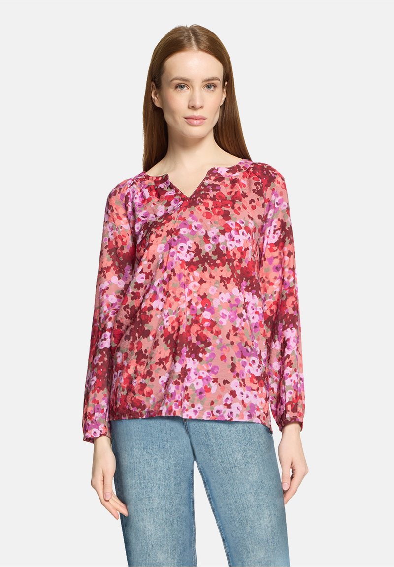 Femme portant un chemisier à manches longues à fleurs dans des nuances de rouge, rose et violet, associé à un jean bleu clair, sur un fond uni.