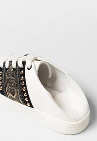 Zapatilla blanca con panel lateral negro que presenta tachuelas doradas y un bordado intrincado, atada con cordones blancos, sobre un fondo simple.