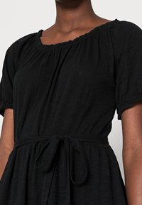 Eine Nahaufnahme einer Person, die ein schwarzes Kleid mit einem sanften Ausschnitt und einer gebundenen Taille trägt, wobei die Textur des Stoffes und die Designdetails hervorgehoben werden.