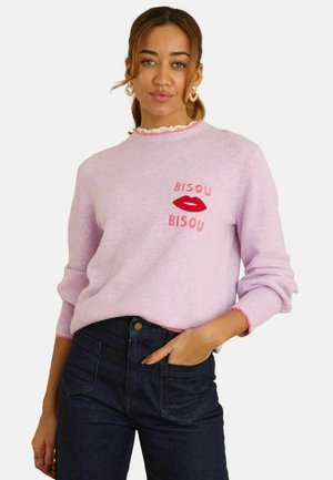 Kvinde iført en lyserød sweater med teksten "BISOU BISOU" og grafik af røde læber, kombineret med mørkeblå jeans, med den ene hånd i lommen.