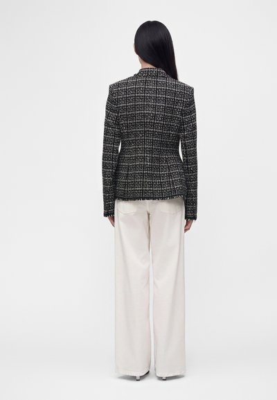 Femme aux longs cheveux foncés portant un blazer à motifs noir et blanc et un pantalon blanc à jambes larges, debout de dos sur un fond uni.