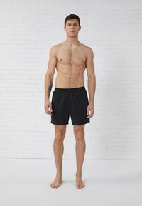 Melni peldkostīmi ar elastīgu jostasvietu, kas atrodas virs ceļgaliem, tos valkā kailkrūts vīrieša modelis pret baltu ķieģeļu sienu.