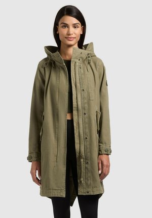 Frau trägt einen olivgrünen Parka mit Kapuze über schwarzer Kleidung, steht vor einem schlichten hellen Hintergrund.