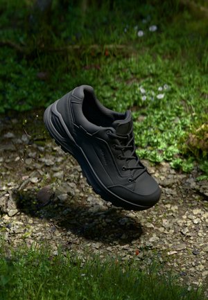 Zwarte hiking schoen met een gestructureerde bovenkant, vetersluiting, stevige zool en een lichtgewicht ontwerp, weergegeven terwijl deze boven een rotsachtige ondergrond zweeft.