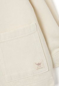 Beige Baumwolljacke mit diagonal gestreifter Textur, einer Klappentasche mit einem kleinen Etikett und sichtbaren Nähdetails.