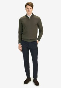 Olivgrön stickad pullover med halvmatchande krage, i kombination med marinblå byxor. Accessoarer inkluderar bruna mockaskor med texturerad tyg.